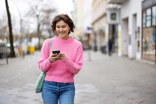 Portrait of smiling young woman using mobile phone while walking outdoors on city street. Pretty young female walking on sidewalk.