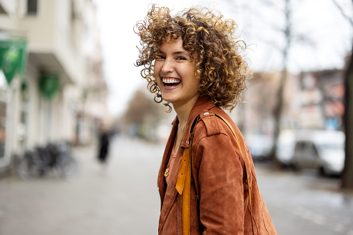 Portrait of cheerful young woman walking outdoors. Female in casual outfit walking on sidewalk looking over the shoulder and laughing at camera.