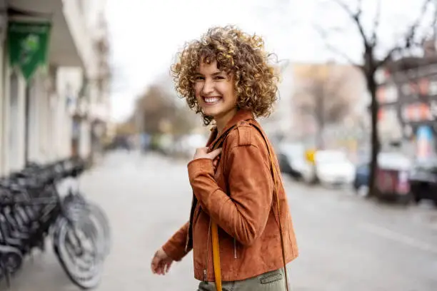 Portrait of smiling woman walking outdoors on city street. Female in casual outfit walking on sidewalk looking over the shoulder.