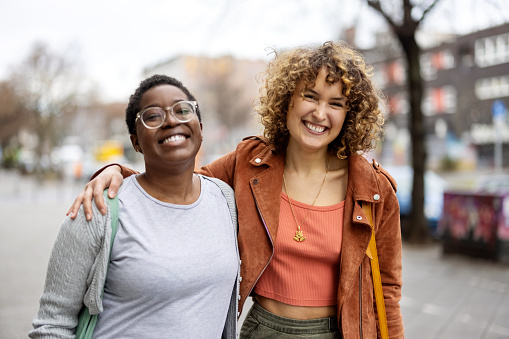 Portrait of two happy females walking together on the street. Diverse women friends in casuals looking at camera and smiling walking outside on the road.