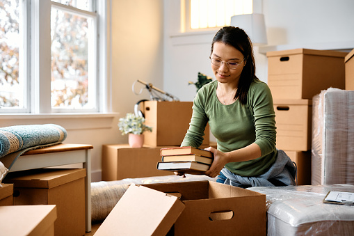 Young Asian woman packing her belongings in cardboard boxes while relocating in new apartment.