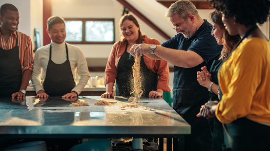 A chef is making pasta from dough in a cooking class with diverse students.