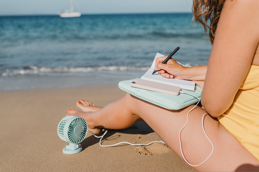 Crop anonymous female tourist taking notes in notepad while sitting on sandy beach with smartphone and portable fan near sea
