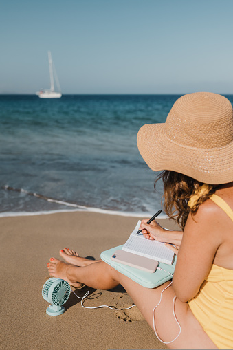 Full body side view of unrecognizable female traveler in hat taking notes in notepad while sitting on sandy coast with fan near sea