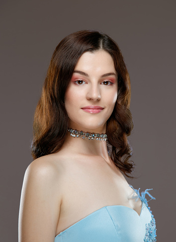 Portrait of beautiful young fashion model with wavy brown hair wearing blue strapless top and necklace, looking at camera and smiling, studio shot, beauty and fashion industry