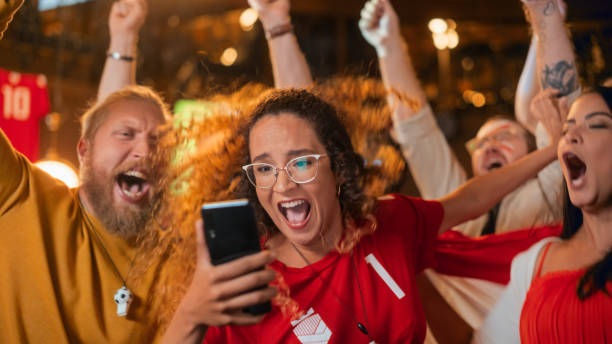 Portrait of an Anxious Multiethnic Female in Red Jersey Holding a Smartphone, Nervous About the Sports Bet on Her Favorite Soccer Team. Happy Victorious Emotions When Football Team Scores a Goal. retrato de una mujer multiétnica ansiosa con una camiseta roja sosteniendo un teléfono inteligente, nerviosa por la apuesta deportiva en su equipo de fútbol favorito. felices emociones victoriosas cuando el equipo de fútbol marca un gol.
