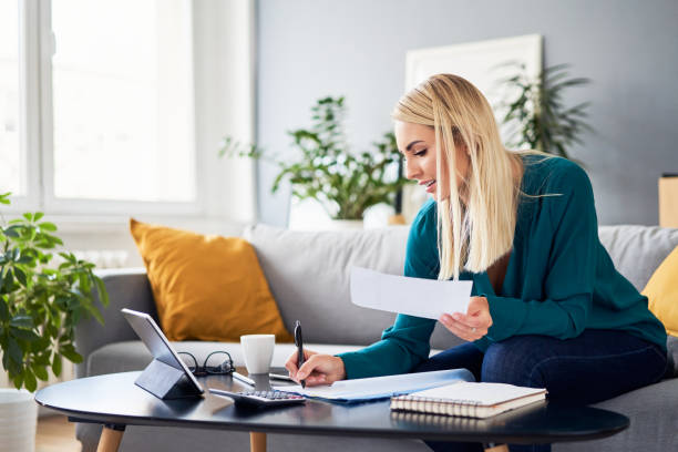 Smiling woman analysing bills filling tax documents while at sitting on sofa at home stock photo