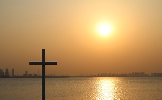 A wooden cross stands atop a platform in front of a tranquil body of water, illuminated by a vibrant sunset