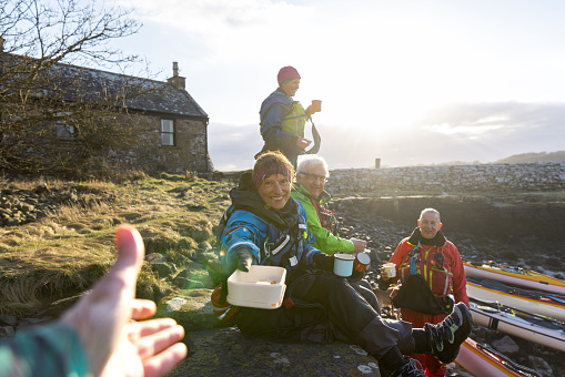 A group of active mature friends enjoying a recreational weekend together in Dumfries, Scotland. They are having a lunch break together and enjoying a hot drink while resting after being on the sea in a kayak. The main focus is a personal perspective of an unrecognisable person receiving a pack lunch box from their friend who is smiling and looking at the camera.