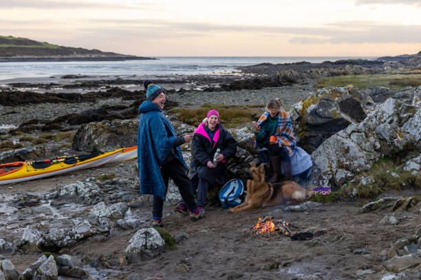 A Happy Life A group of friends enjoying a recreational weekend together in Dumfries, Scotland. They are having a rest on the beach with their dog in warm clothing and blankets after being in the sea. They are all relaxing and laughing around a campfire that is dying out. beach fire scotland stock pictures, royalty-free photos & images