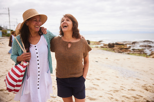Two carefree mature female friends laughing while walking arm in arm together along a sandy beach on an overcast day