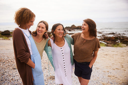 Carefree group of mature female friends laughing while standing arm in arm together on a sandy beach on an overcast day