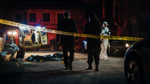 Cinematic Shot: Lieutenant Arriving At a Crime Scene, Crossing the Yellow Tape, Listening to Briefing from First Responder Officer. Detective Checking the Body Bag. Forensics Team Gathering Evidence stock photo