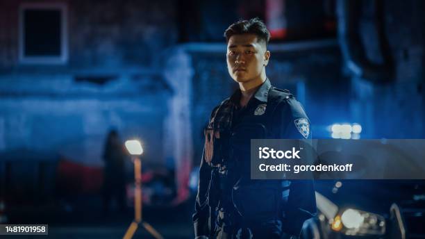 Medium Shot Portrait Of A Young Asian Male Police Officer Looking Away Then Turning To Camera Under Siren Lights Brave Officer Of The Law Keeping Citizens And Civilians Safe Fighting Crime Stock Photo - Download Image Now