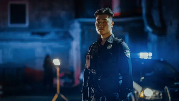 Photo of Medium Shot: Portrait of a Young Asian Male Police Officer Looking Away then Turning to camera Under Siren Lights. Brave Officer of the Law, Keeping Citizens and Civilians Safe, Fighting Crime