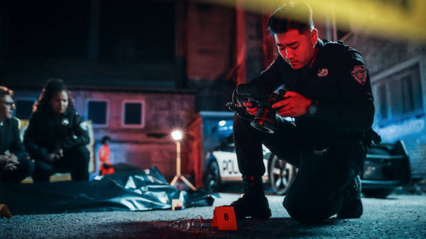 policeman taking photos of marked evidence on a crime scene at night. forensics police officer finds glasses belonging to the killer. detectives talking about the victim's corpse in the background - forensic science flash imagens e fotografias de stock