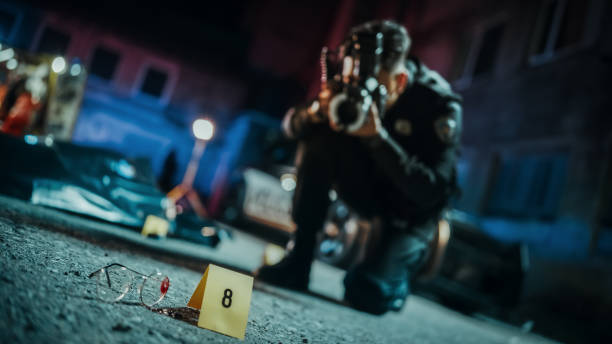 policeman taking photos of marked evidence on a crime scene at night. forensics police officer finds glasses potentially belonging to the dead victim and photographs it for analysis. focus on glasses - forensic science flash imagens e fotografias de stock