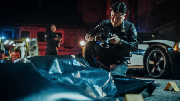 法医学作業を行い、マークされた証拠の写真を撮るアジアの若い警察官の広角ポートレートショット。救急隊員とバックグラウンドで協力する犯罪現場調査ユニット - forensic science flash ストックフォトと画像