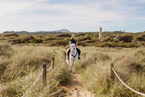 Beautiful young blond woman riding out with her white horse between the green sand dunes. Color editing. Part of a series.