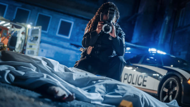 black female police officer using camera with flash to take photos of a dead body covered by a cloth at a crime scene. policewoman professionally documenting evidence for the case file - forensic science flash imagens e fotografias de stock