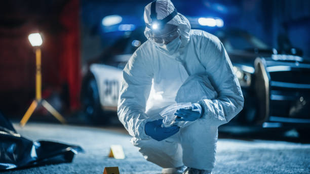 istock Portrait of Two Forensics Experts Doing Fieldwork at Night at a Crime Scene. One Technician Packs the Bloodied Knife as Murder Weapon While the Other is Taking Photos of the Dead Body. 1481911531
