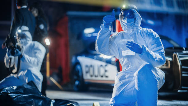 カバーオールスーツを着た2人の男性スペシャリストが犯罪現場で証拠を収集しています。ある専門家は、滅菌されたビニール袋にナイフを入れています。2つ目は、被害者の袋詰めされた遺� - forensic science flash ストックフォトと画像