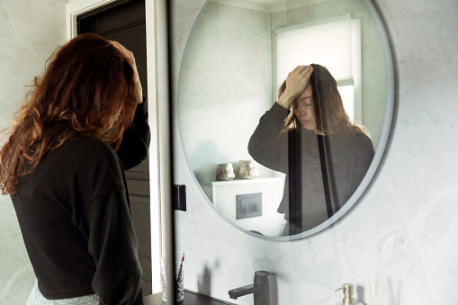Depressed Woman holding head and looking down in front of bathroom Mirror