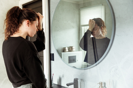 Depressed Woman holding head and looking down in front of bathroom Mirror