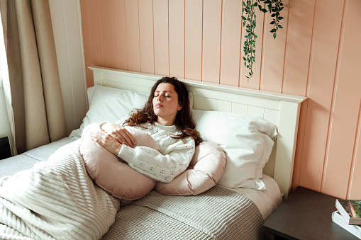 Young european woman in painful expression holding her belly suffering menstrual period pain lying sad on home bed having tummy cramp in female health concept