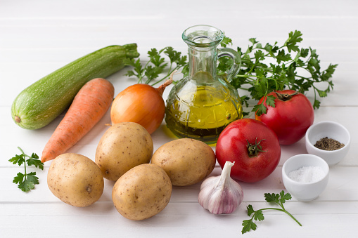 Ingredients for vegetable stew: potatoes, zucchini, tomatoes, onions, garlic, parsley, olive oil, salt and black pepper on a white wooden background
