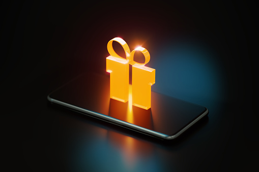 Yellow gift symbol glowing over a smart phone on black background. Horizontal composition with copy space.