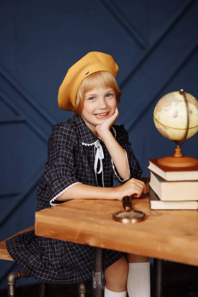 A little schoolgirl in a plaid school uniform and a French beret is sitting at her desk with a serious face. The concept of studying geography, elementary school, information search.