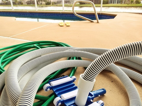 Pool vacuum attachment and hoses for cleaning a pool. Pool season brings pool chores to starting the season. Pool deck, sandals and edge of pool shown in blurred background with copy space.