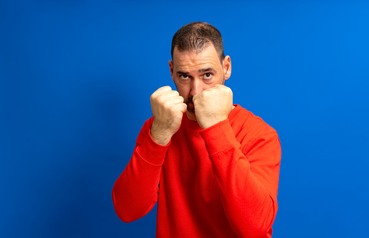 Bearded Hispanic man in his 40s wearing a red jersey standing guard ready to start a fight with his fists, isolated over blue background