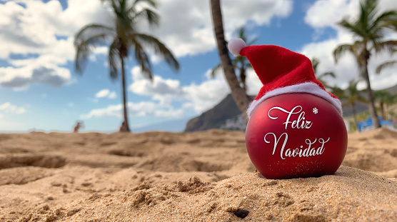 Christmas bomb in Santa's hat with words Merry Christmas in spanish on the beach lying on the sand with palm trees and blue sky on the background.