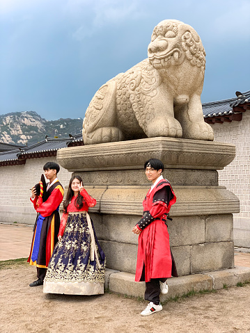 Three young South Koreans dressed in traditional costumes pose with Haetae (Haechi) guardian lion at Gwanghwamun gate in Seoul. Girl in the middle smiling at camera, striking a V sign. Photo taken on public road.