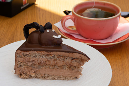 White plate with chocolate piece of cake with cream and edible chocolate mouse on top of it beside of cup of green tea.