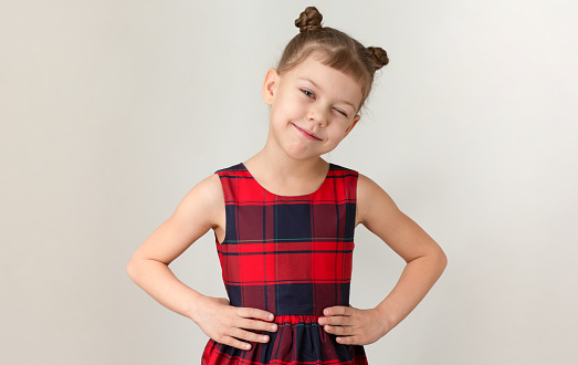 Winking caucasian kid child little girl of 6 7 years in red plaid dress holding hands on waist on grey background looking at camera
