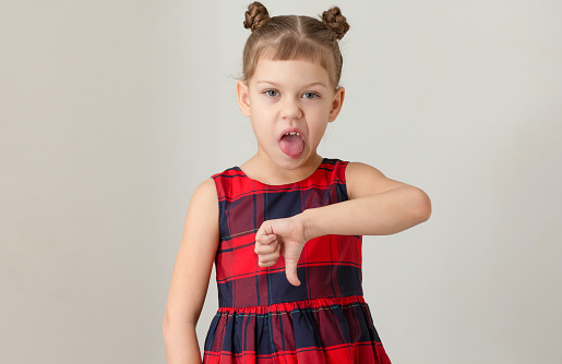 Child giving thumb down with funny face expressing disgust with open mouth and showing tongue caucasian 6 7 years in red plaid dress
