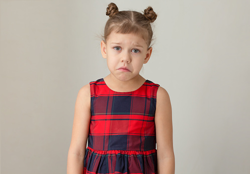 Sad and unhappy child because of distress, pain or sorrow on grey background caucasian kid little girl of 6 7 years in red plaid dress