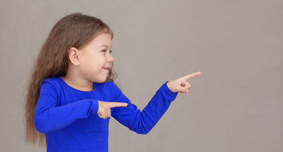 Smiling and happy little girl showing by index fingers on two hands on copy space isolated on white background, looking aside waist up caucasian little girl of 5 years in blue