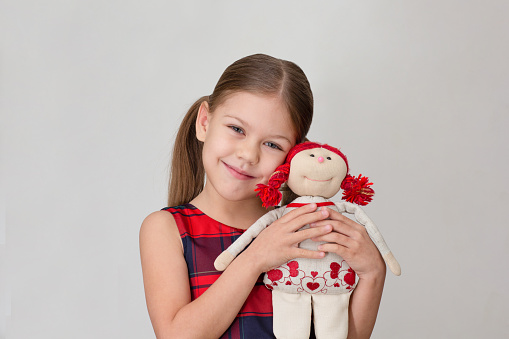 Happy and smiling child hugging doll toy on grey background caucasian little girl kid of 6 7 years in red plaid dress looking at camera