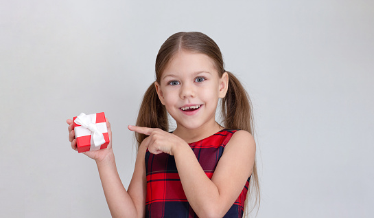 Happy and smiling child holding little box as present and showing on it by forefinger on grey background, caucasian little girl kid of 6 7 years in red plaid dress looking at camera