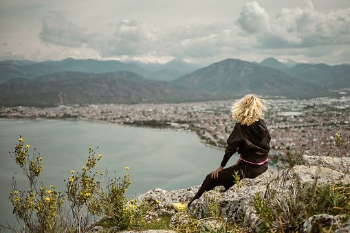 A blonde woman sits on a high rock overlooking the seaside town and bay in the Aegean Sea