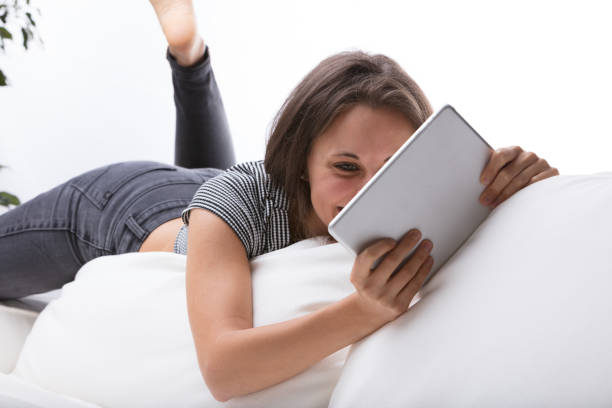 Young woman lounging on the edge of the backrest of a white couch, lazily contorted because of her fit body, looks at something on the invisible screen of a digital tablet - maybe a video, a book or a