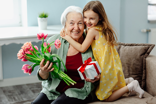 Happy granddaughter and grandmother laugh at the original congratulations on the holiday. A smiling caring granddaughter presents flowers and a handmade gift to an elderly grandmother on Women's Day.