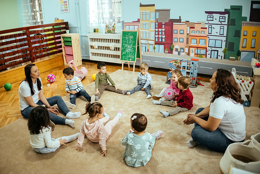 A small group of Kindergarten students sit on the floor with their teacher as they each hold a set of bells sing along together.  They are each dressed casually and are focused on shaking their instruments to the beat.