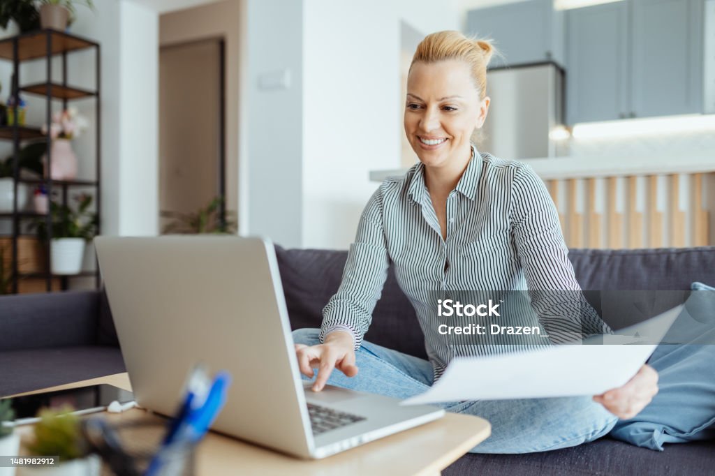 Mature entrepreneur working from home on laptop, anaylzing documents and doing paperwork Female entrepreneur or freelance worker doing business from couch Internet Stock Photo