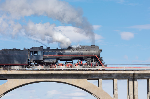 A smoking steam locomotive driving over an old bridge against a blue sky