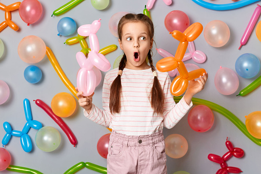 Funny shocked surprised little girl with pigtails standing against gray wall decorated with colorful balloons, looking away, sees surprise, excellent birthday present.